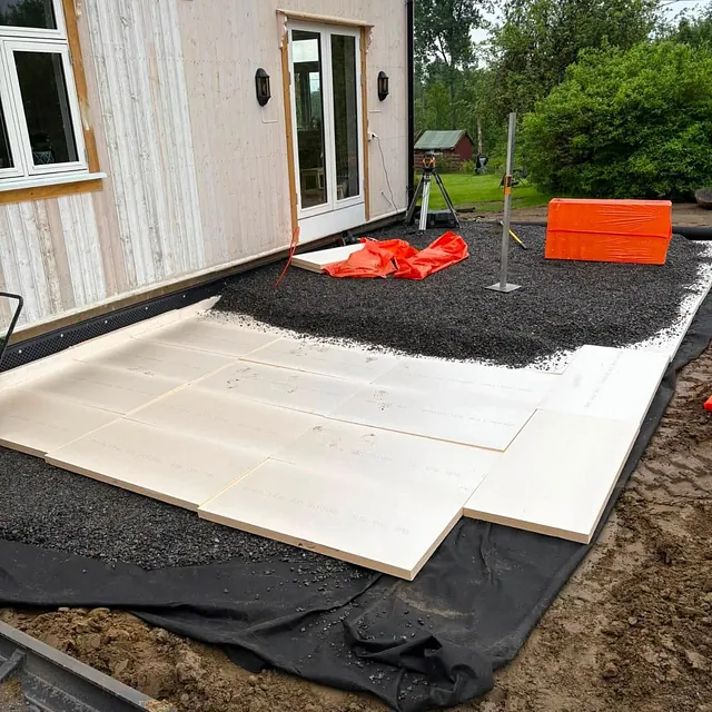Isolasjonsplater og duk lagt i bakken langs hus med lys kledning